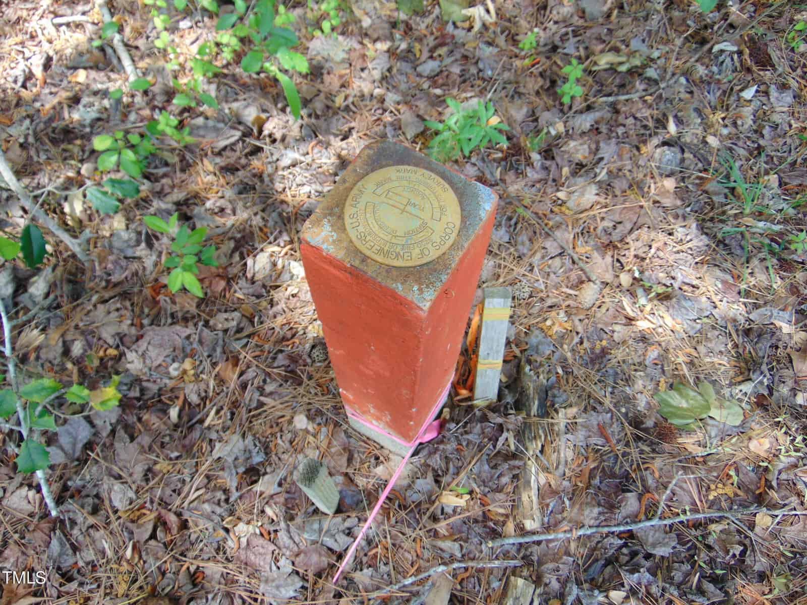 A rectangular red concrete survey marker stands in a wooded area with pine needles and leaves on the ground; a circular brass plaque with text is affixed to the top. A small orange and white post is next to it.