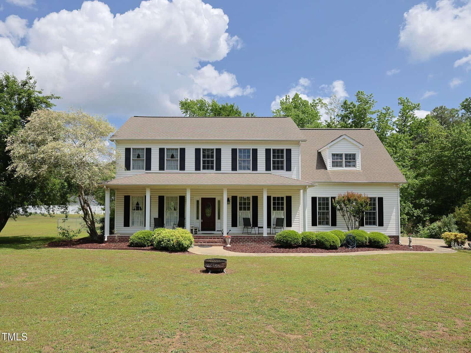A large, two-story white house with black shutters, a front porch, and neatly trimmed bushes, set on a spacious grassy lawn with trees and a blue sky in the background.