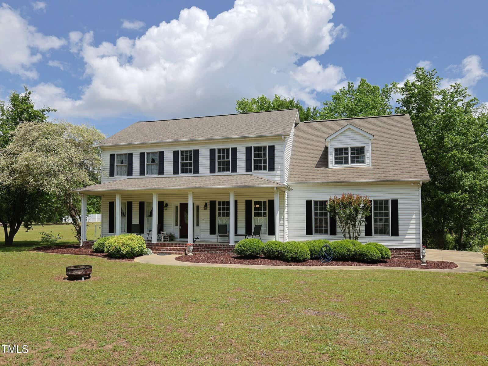 A large two-story white house with black shutters, a covered front porch supported by columns, surrounded by green lawn, shrubs, and trees under a partly cloudy sky.