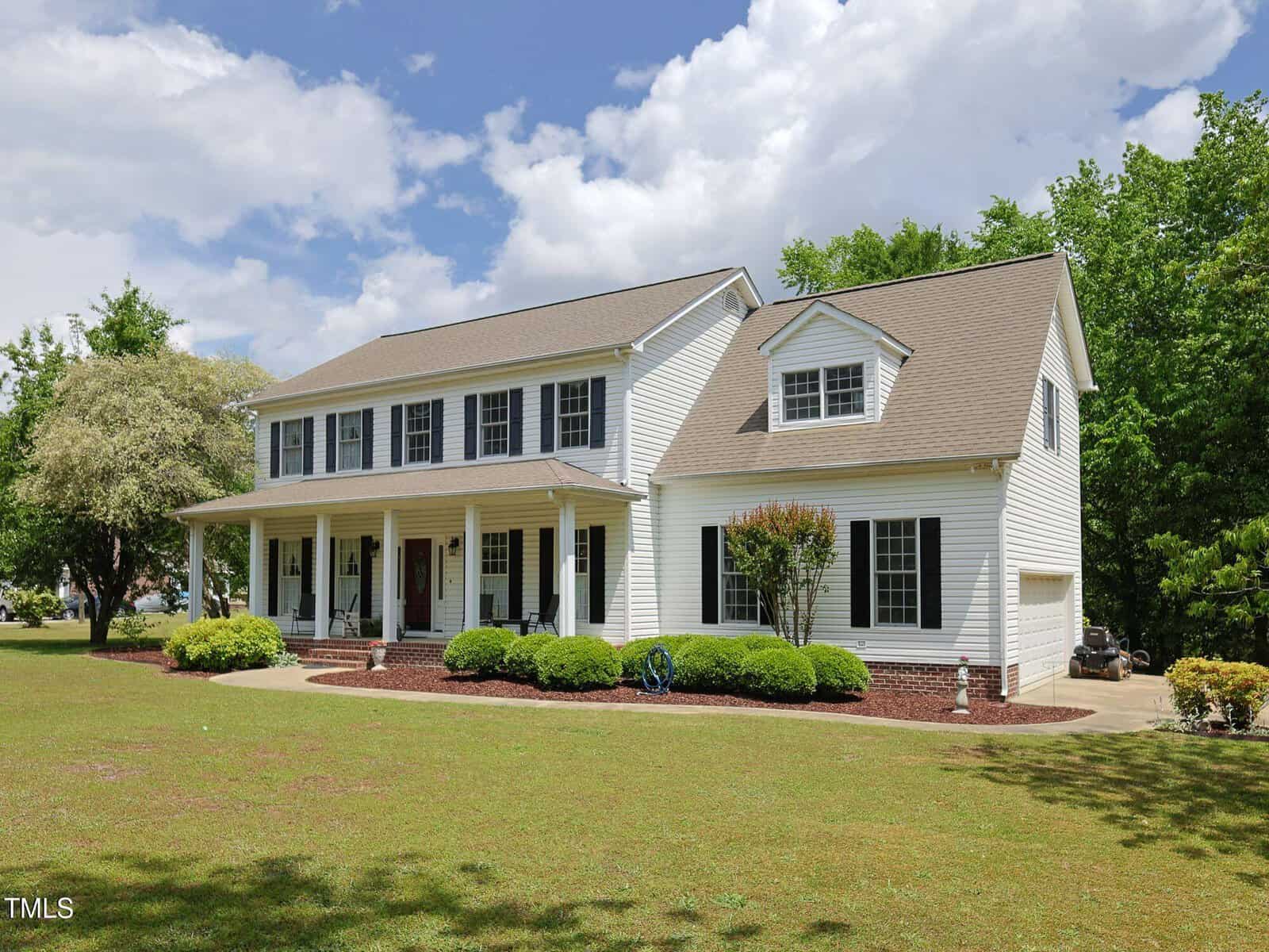 A large two-story white house with black shutters, a covered front porch supported by columns, and a well-maintained lawn with shrubs and trees under a partly cloudy sky.