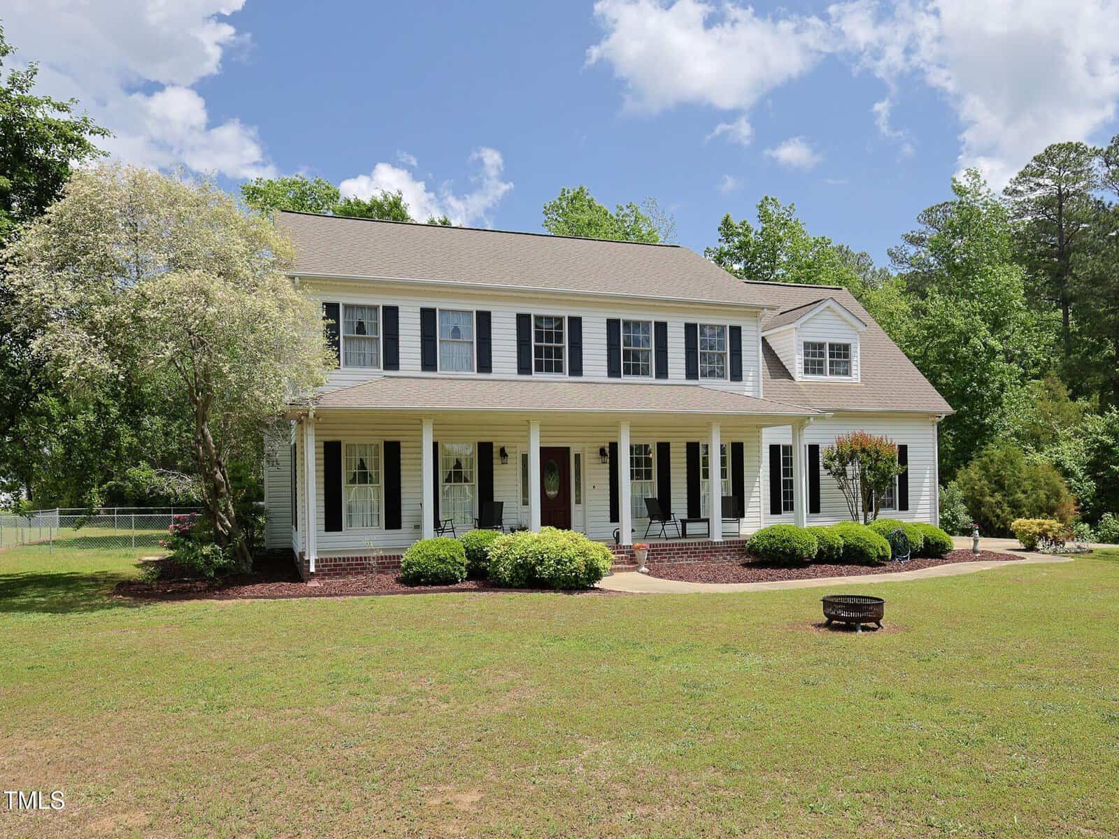 Large two-story white house with black shutters, a covered front porch with chairs, surrounded by shrubs and trees, and a spacious green lawn under a partly cloudy sky.