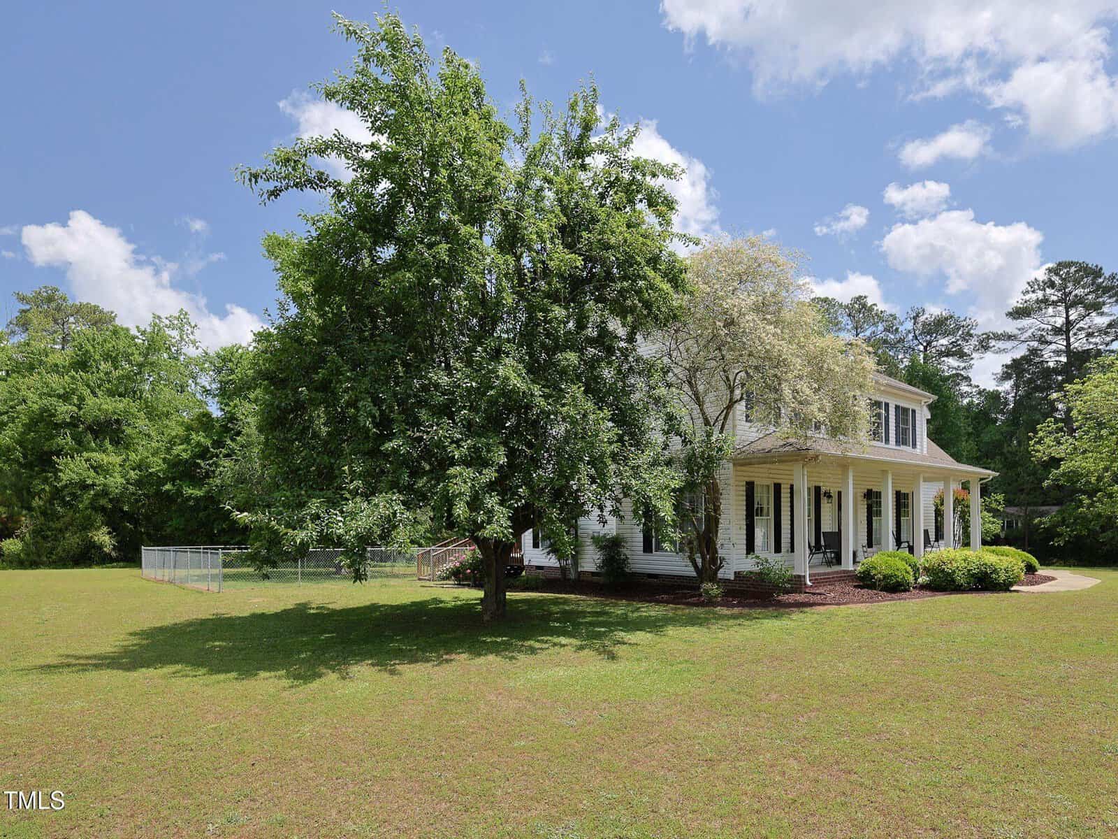 A white two-story house with a covered porch is partially obscured by large trees in the front yard. The house sits on a grassy lot with a fenced area to the left and is surrounded by greenery under a blue sky.