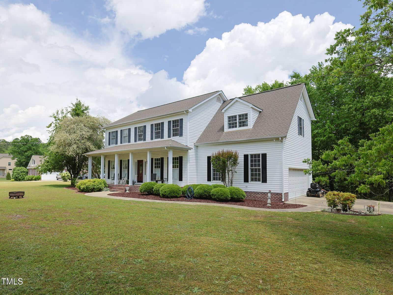 A large two-story white house with a covered front porch, multiple windows, and a neatly landscaped yard. The house sits on a spacious grassy lawn bordered by trees under a partly cloudy sky.