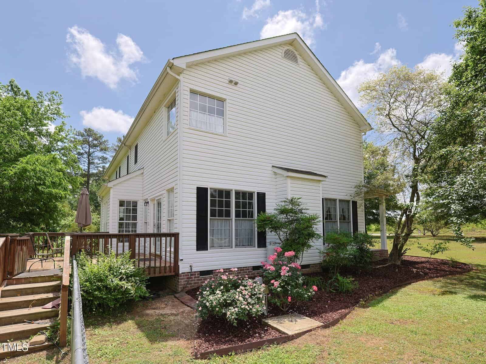 Two-story white house with black shutters and a wooden deck on the side. There are bushes and flowering plants along the house, and a sloped yard with lawn and trees under a partly cloudy sky.