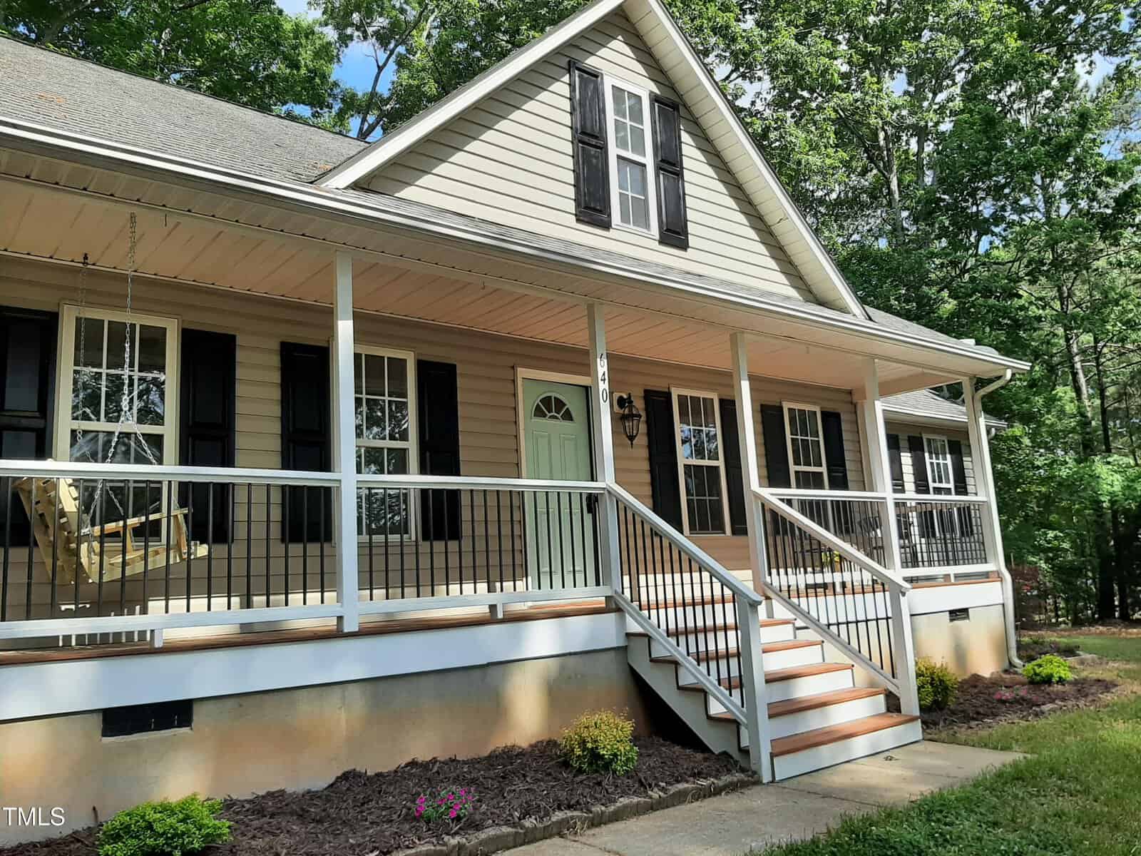 A beige house with a wide front porch, black shutters, a mint green door, white railings, a porch swing, and steps leading up. The house is surrounded by green trees and small landscaped bushes.