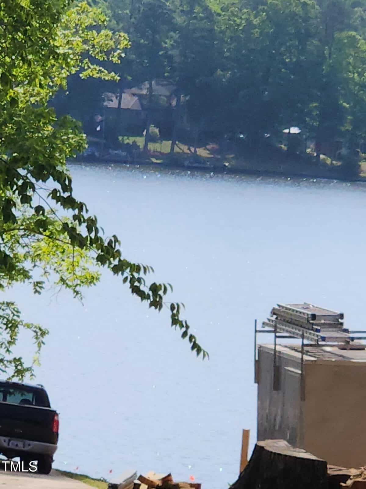 View of a calm lake bordered by trees, with a few houses visible on the far shore. In the foreground, there is a partially constructed building, a parked truck, and stacked construction materials.