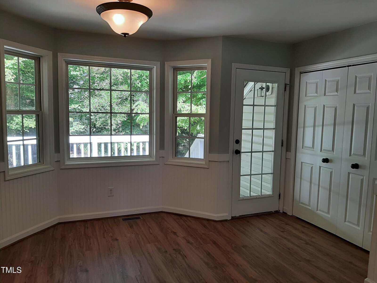 A bright, empty room with three large windows, a glass door, and white double closet doors. The room has light gray walls, a ceiling light fixture, and wood floors. Trees and a porch are visible outside.