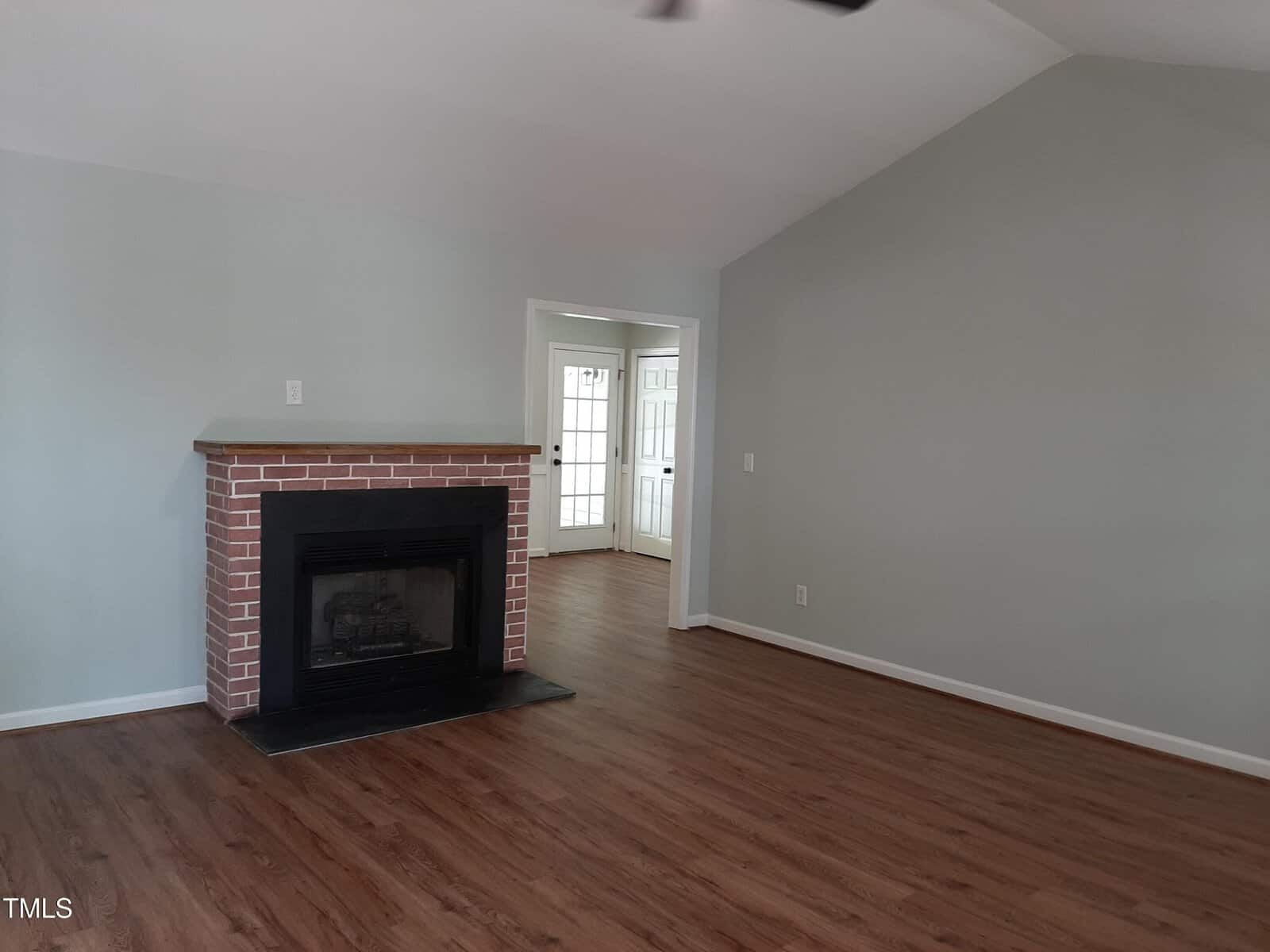 A spacious living room with light gray walls, wood flooring, a brick fireplace, and an open doorway leading to a bright room with French doors.