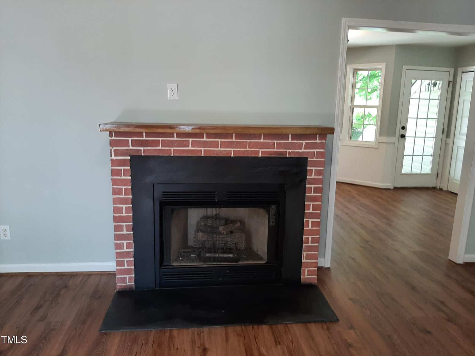 A brick fireplace with a black metal frame stands against a gray wall. The room has wood flooring, and an adjoining room with large windows and a white door is visible in the background.