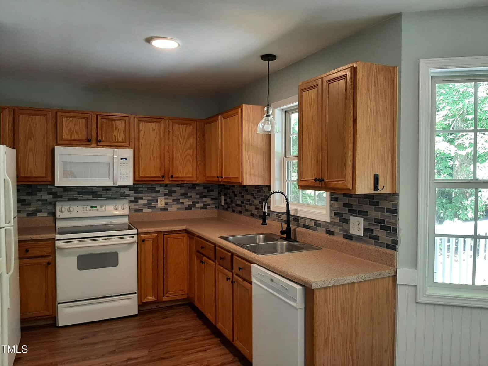 Kitchen with wooden cabinets, white appliances including oven, microwave, and dishwasher, double sink, tiled backsplash in gray and blue tones, and a window providing natural light. Countertops are beige and flooring is wood.