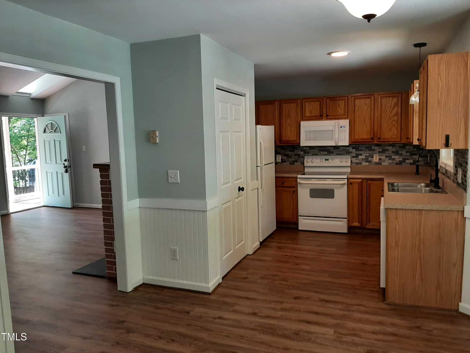 Kitchen with wood cabinets, white appliances, tile backsplash, and laminate flooring. An open doorway leads to a bright living area with large windows and a brick half wall. Walls are painted light gray and white.