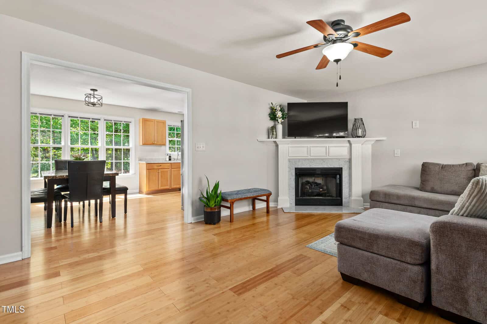 A living room with light wood floors, a gray sectional sofa, a white fireplace with a TV on top, a ceiling fan, a potted plant, and an adjoining dining area with large windows in the background.