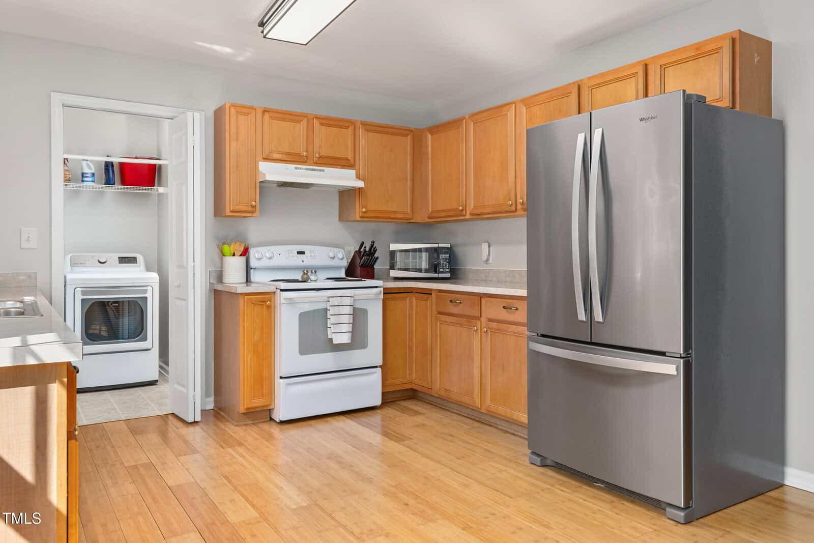 A kitchen with light wood cabinets, white appliances including a stove and range hood, and a stainless steel French door refrigerator. A laundry room with a washer and dryer is visible through an open door.