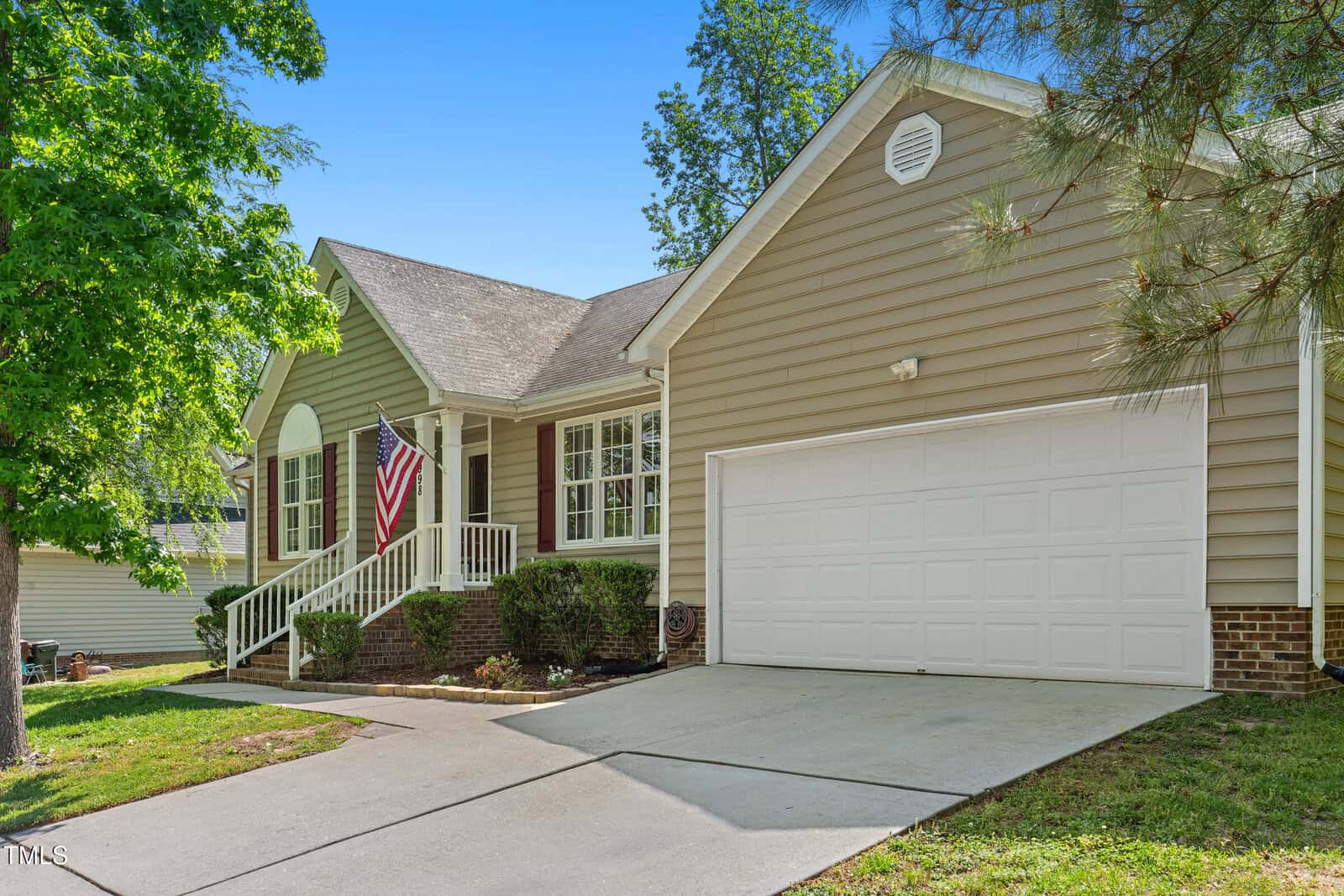 A beige single-story house with white trim, a two-car garage, a front porch with railing, an American flag, and a sloped driveway. There are bushes along the porch and trees around the yard.
