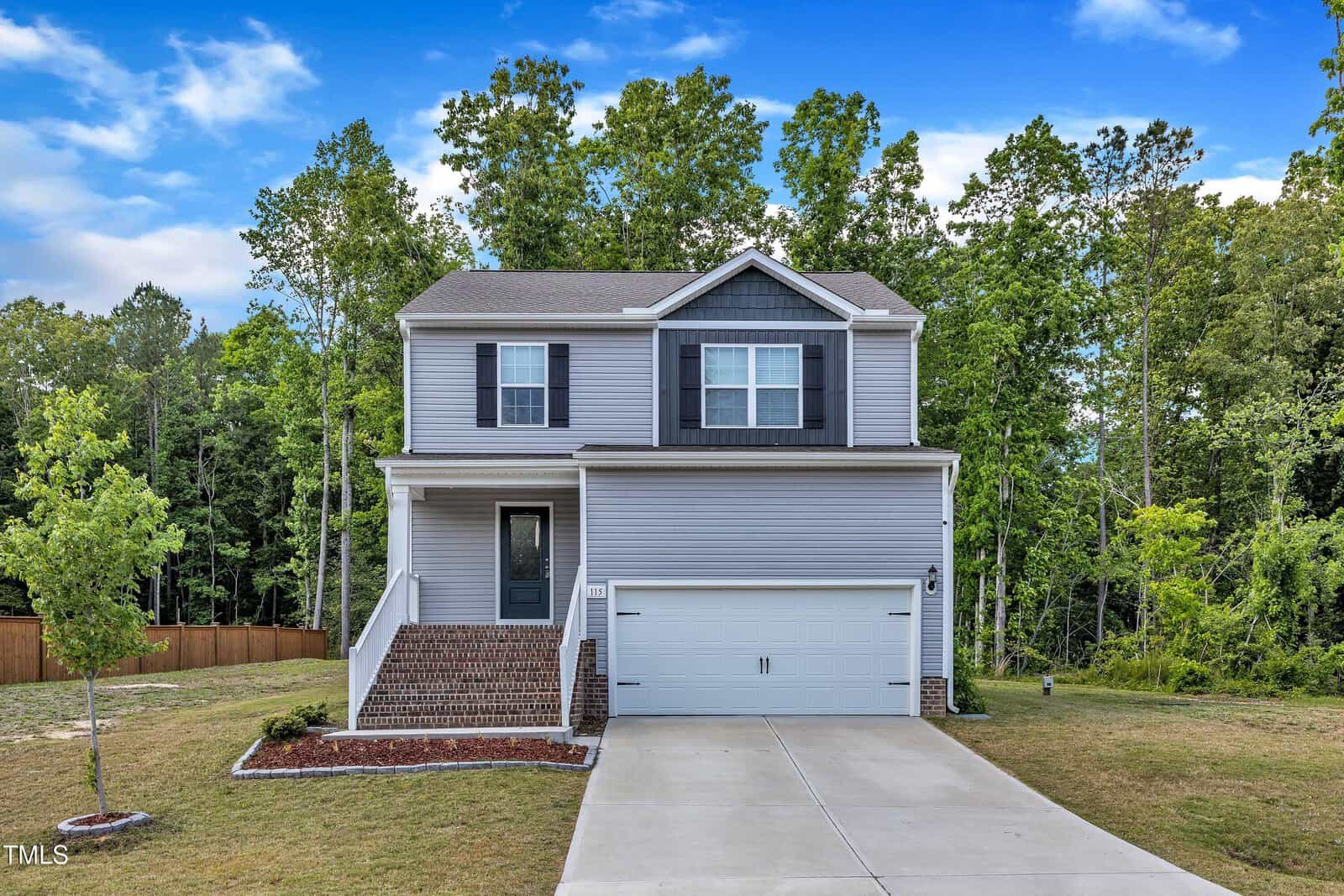 Charming two-story gray house with white trim at 115 Parkers Gate Drive in Youngsville, featuring a covered front porch, brick steps, attached two-car garage, and a concrete driveway. The home is surrounded by lush grass, a few small trees, and a wooded area in the background.