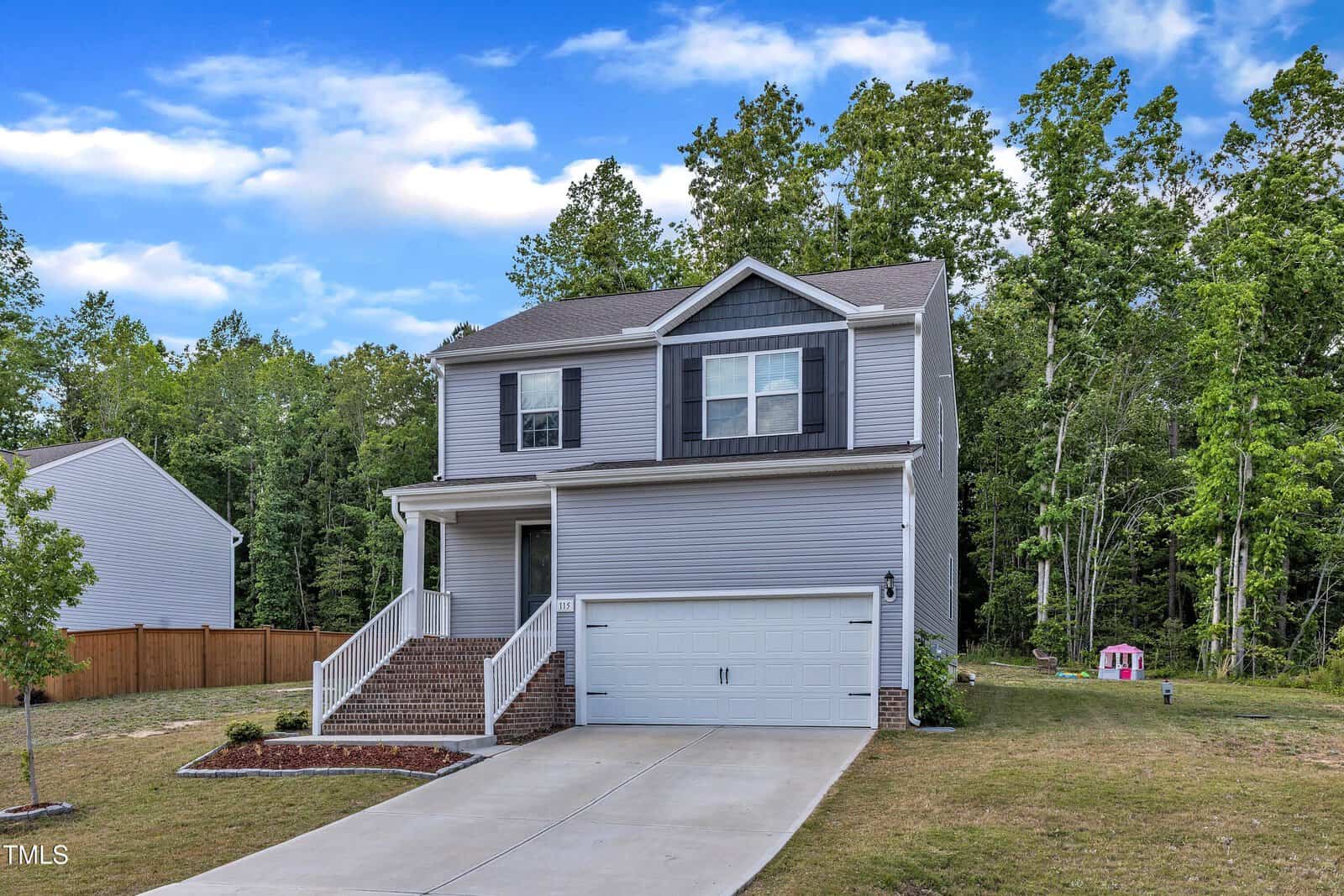 Two-story gray house with white trim, covered front porch, attached two-car garage, and a sloped driveway. The yard is grassy with a small tree and a pink playhouse near the wooded area in the background.