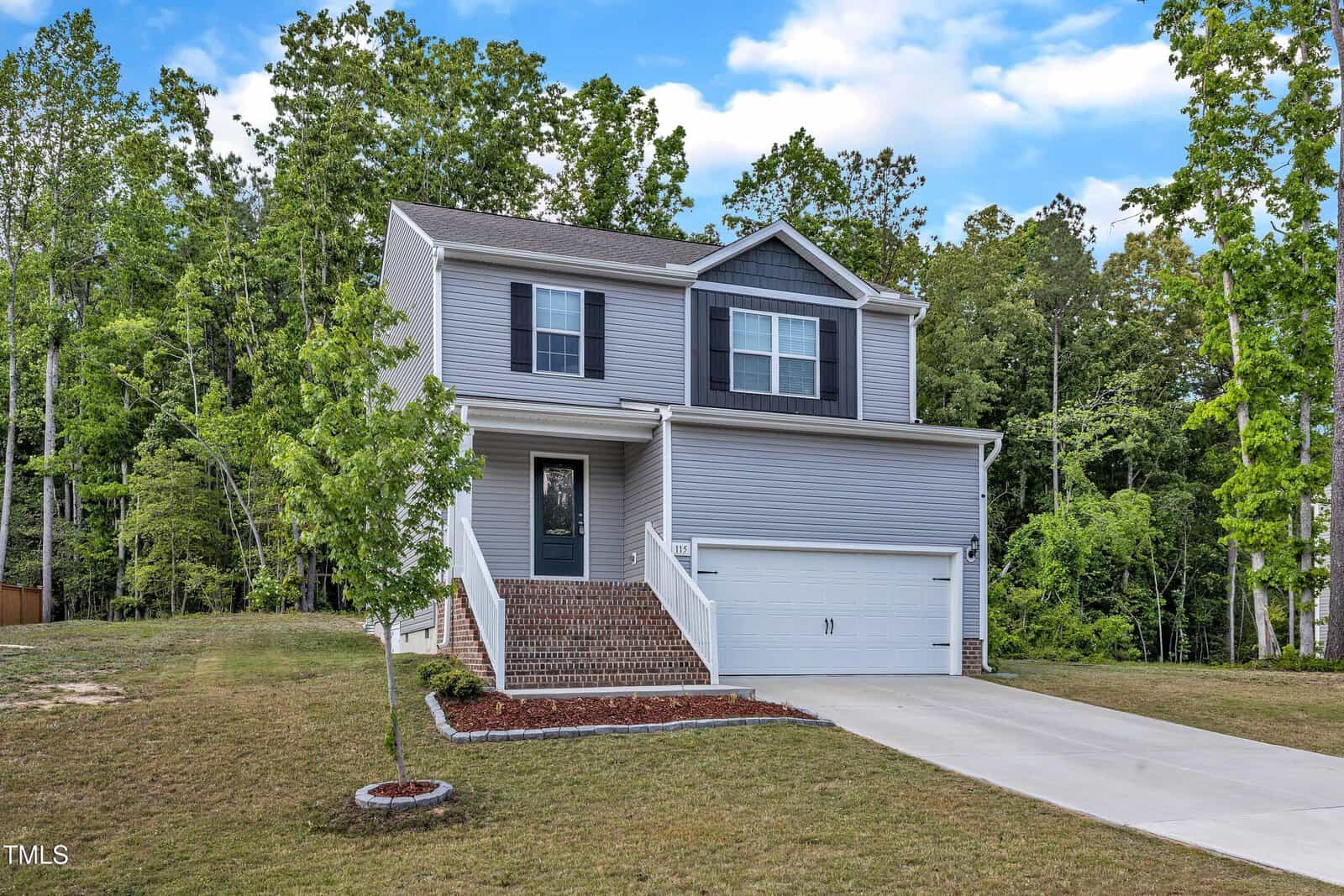 Two-story gray house with white trim, an attached two-car garage, front steps leading to the entrance, a small front yard with minimal landscaping, and surrounded by tall trees under a partly cloudy sky.