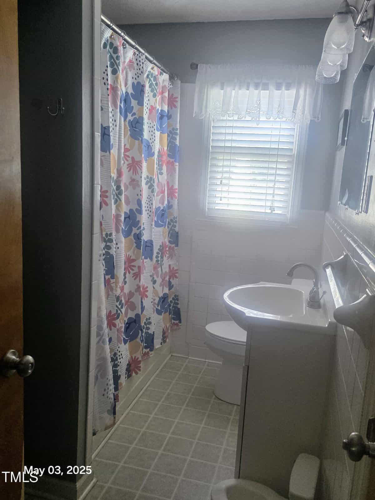 A small bathroom with a shower and a colorful floral curtain, a white toilet, a white sink with a cabinet, tiled walls, a window with blinds and a lace curtain, and a tiled floor. A timestamp reads "May 03, 2025.