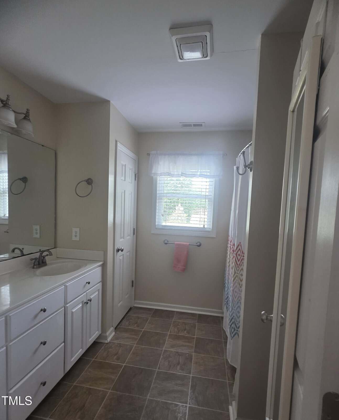 A bathroom with beige walls, a white double-sink vanity with a mirror, tiled floor, a window with a sheer curtain, a towel bar with a pink towel, a shower with a patterned curtain, and a ceiling vent.
