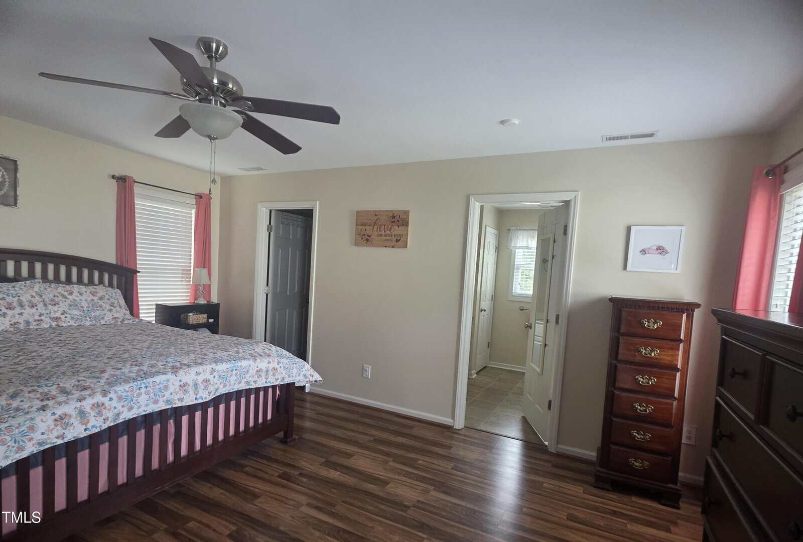 A bedroom with a wooden bed, floral bedding, two windows with pink curtains, a ceiling fan, two dressers, and a doorway leading to a bathroom. The floor is wood and walls are light-colored.