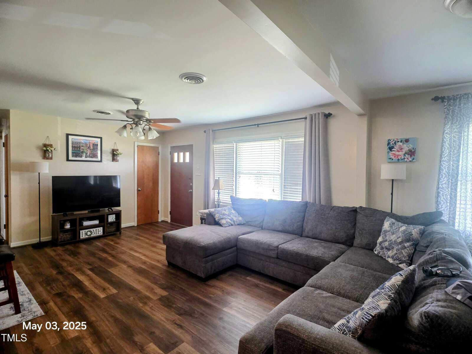 A living room with a large gray sectional sofa, a TV on a stand, wood flooring, ceiling fan, and windows with blinds and curtains. A door and framed art are visible; the photo is dated May 03, 2025.