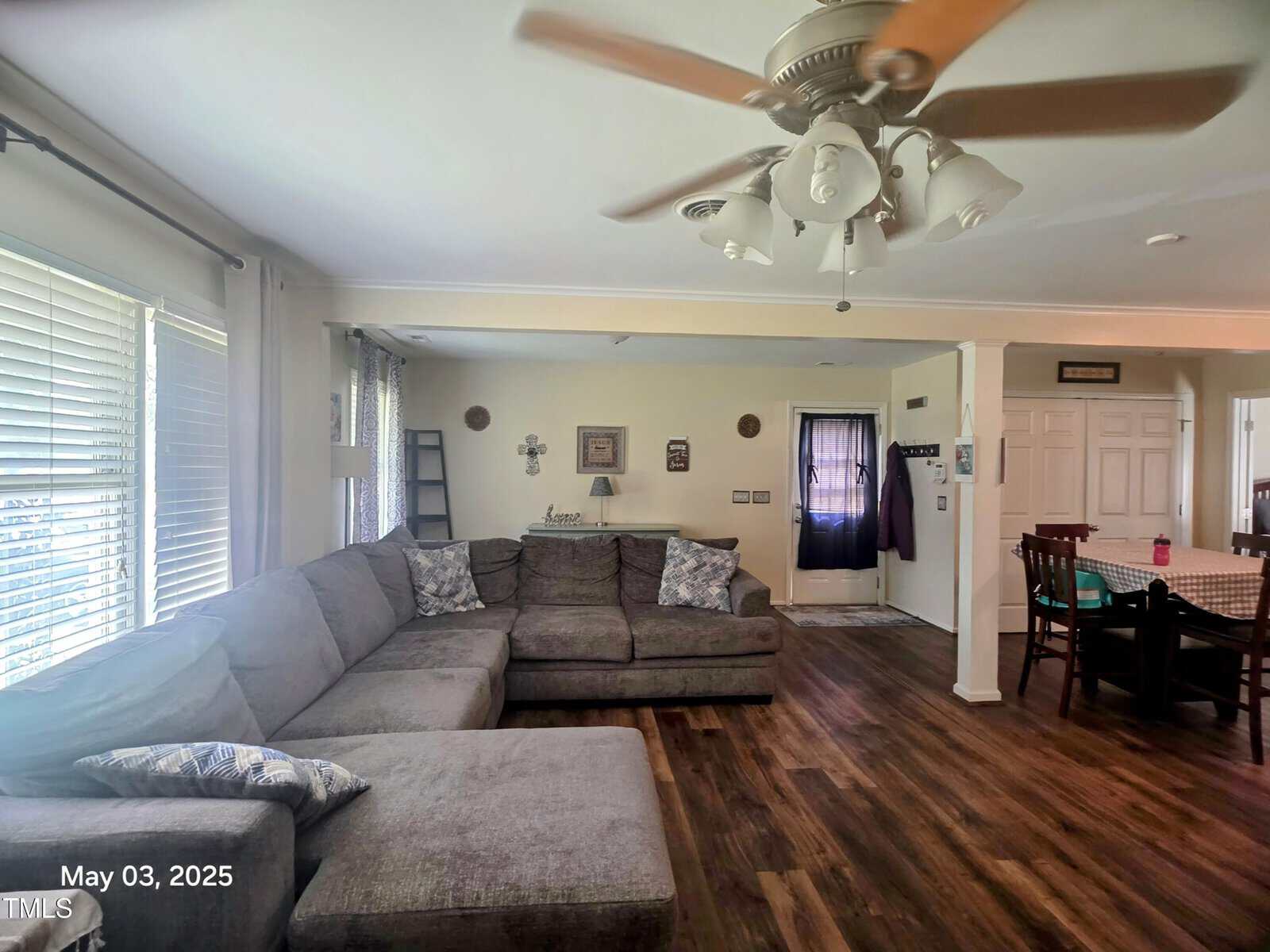A living room with a large gray sectional sofa, wooden floor, ceiling fan, and various wall decorations. Sunlight streams through wide windows on the left. Part of a dining area and entryway are visible in the background.