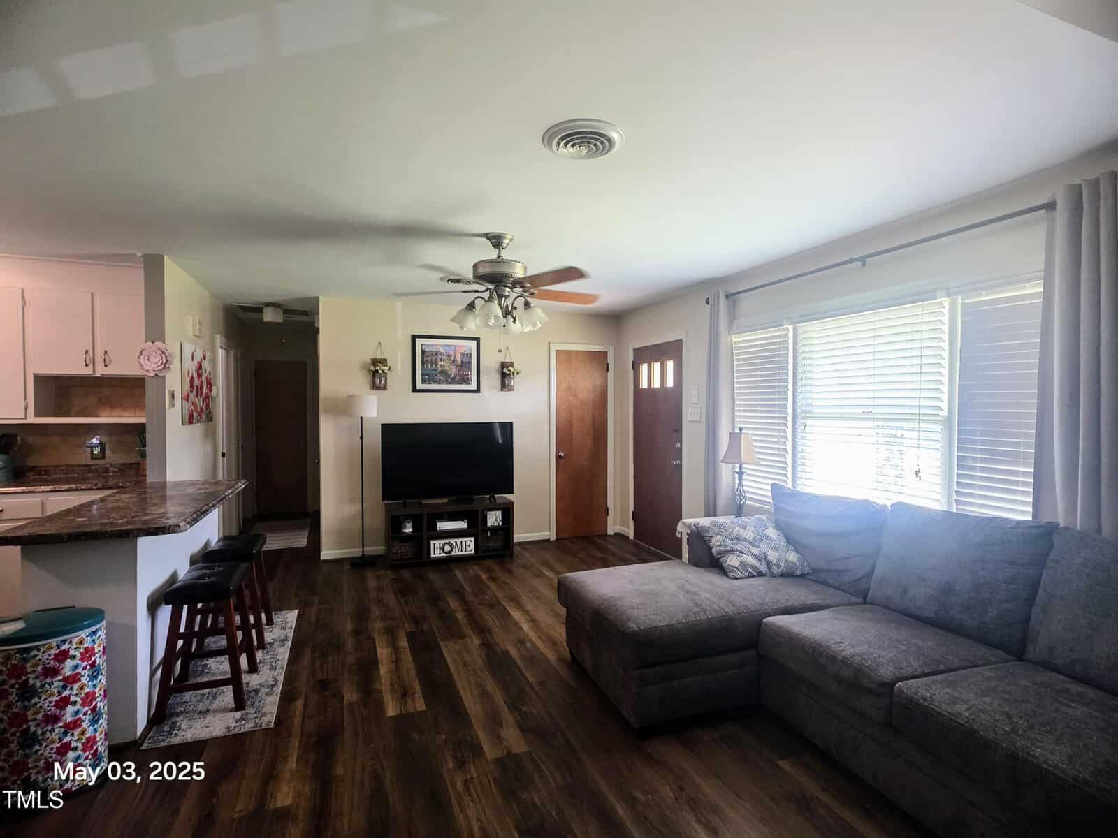A living room with wood flooring, a gray sectional sofa, a TV on a stand, bar stools at a kitchen counter, a ceiling fan with lights, and large windows with blinds. The date "May 03, 2025" appears in the corner.