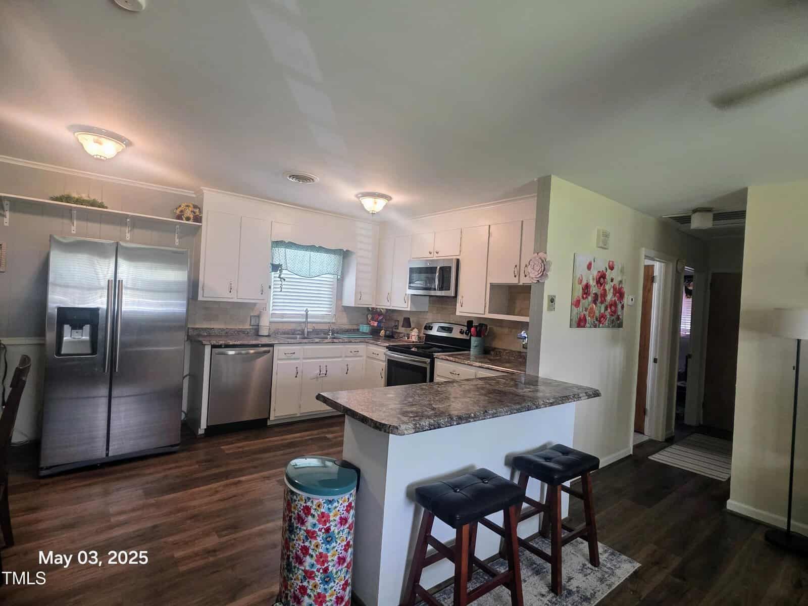 A modern kitchen with white cabinets, stainless steel appliances, an island with two stools, a trash can, wood flooring, and decorative wall art. The date May 03, 2025, appears in the bottom corner.