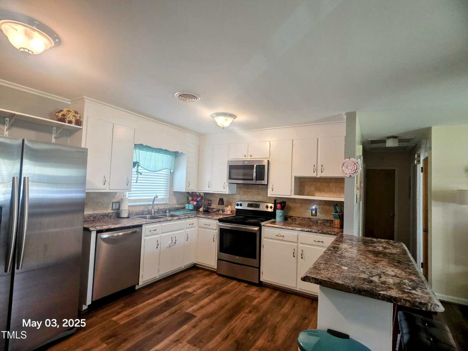A kitchen with white cabinets, stainless steel appliances, brown patterned countertops, a window with a blue curtain, and wood-look flooring. The date "May 03, 2025" is visible in the bottom left corner.