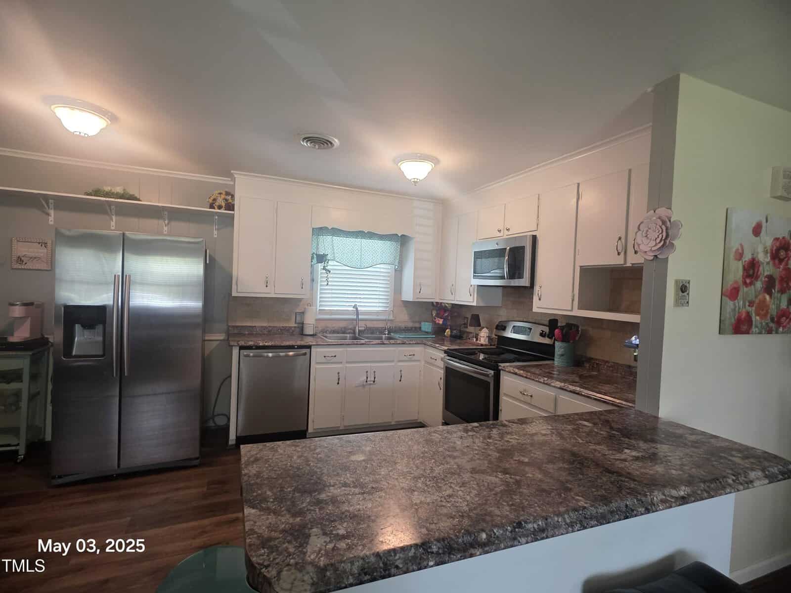 A kitchen with white cabinets, stainless steel appliances, a double sink under a window with a curtain, a brown speckled countertop, and a breakfast bar in the foreground. Date in corner reads May 03, 2025.