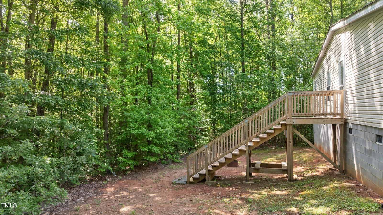 A wooden staircase leads from the side entrance of a house down to a dirt yard bordered by a dense forest of green trees. The house has light-colored siding and a block foundation.