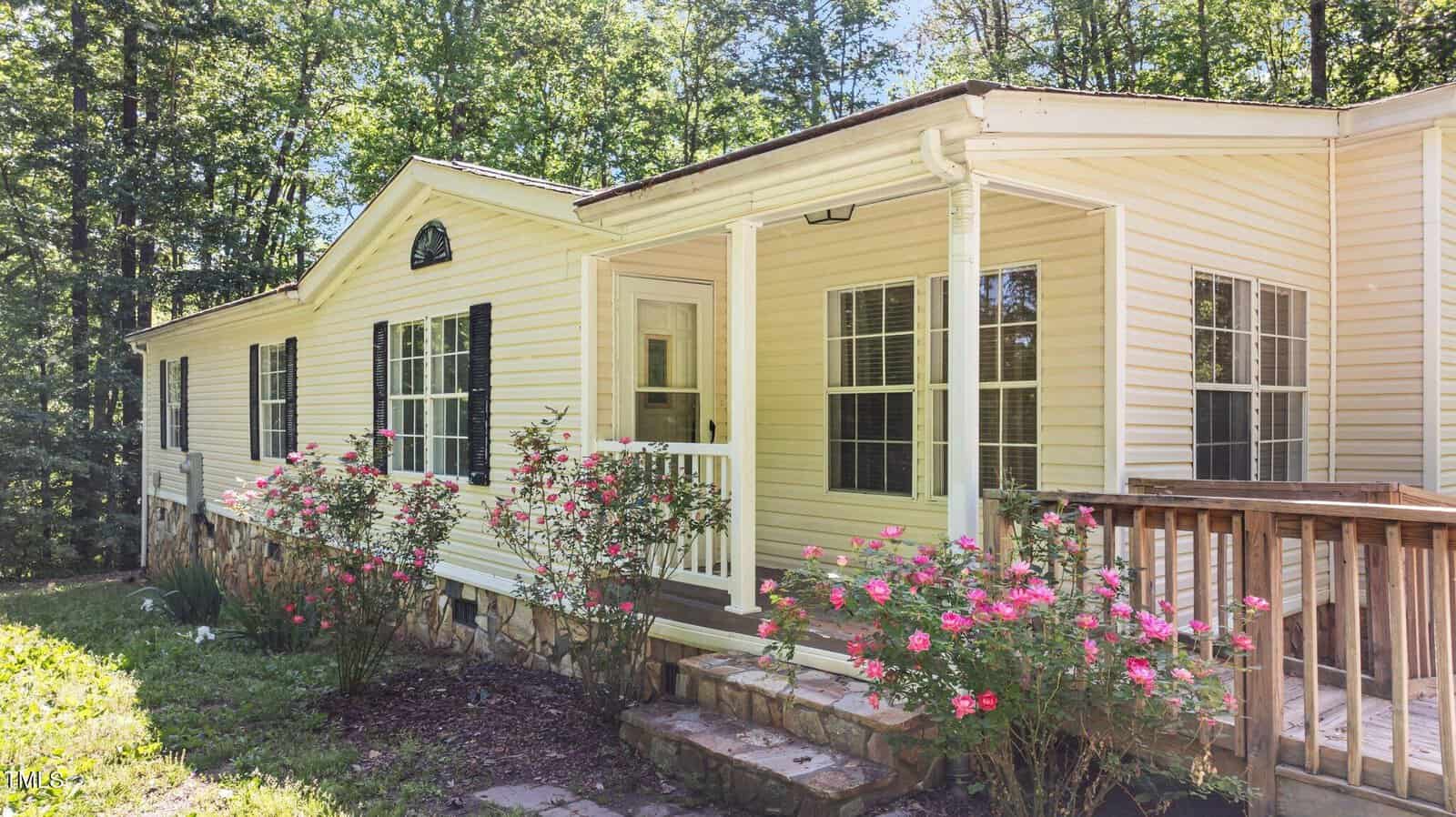 A single-story light yellow house with white trim features a small covered porch, black shutters, and blooming rose bushes along the stone-bordered foundation, set against a backdrop of tall trees.