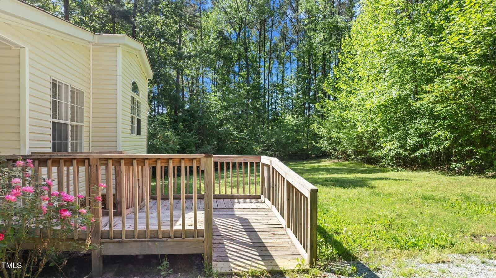 A small wooden deck attached to a yellow house overlooks a grassy yard bordered by dense green trees. Pink flowers are growing beside the deck, and sunlight filters through the trees in the background.