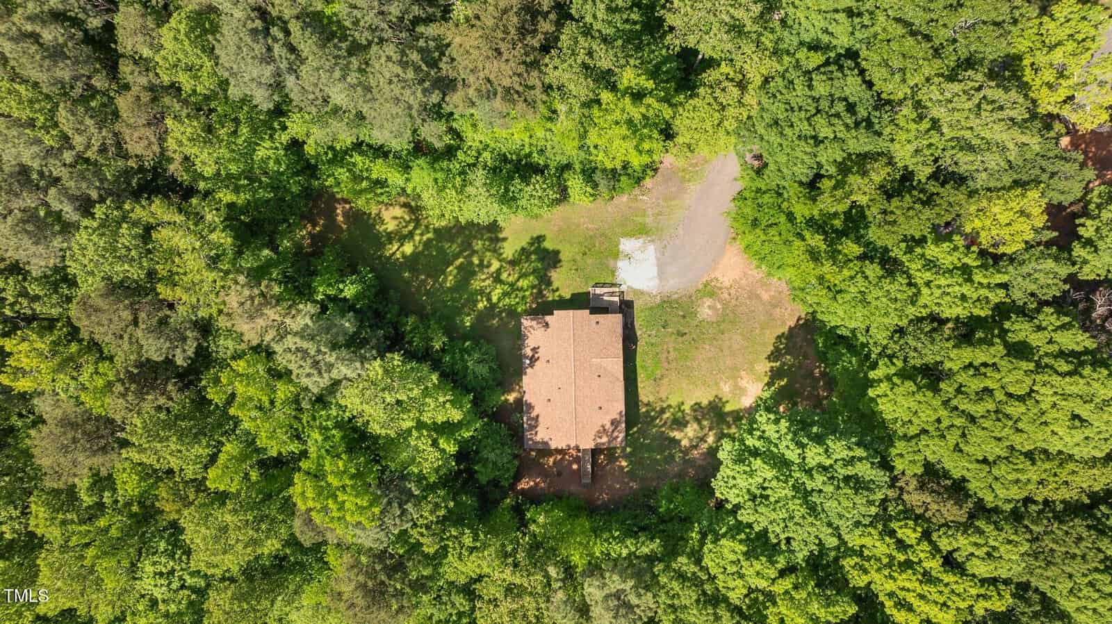 Aerial view of a single-story house with a tan roof surrounded by dense green trees, with a small clearing and driveway visible next to the house.