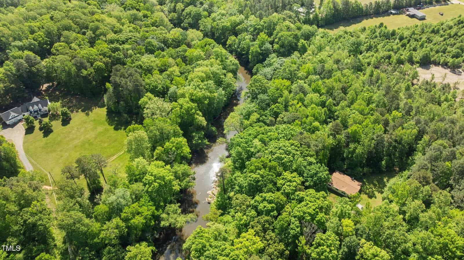 Aerial view of a forested area with a river running through the center, a house with a driveway on the left, and another house partially visible among trees on the right. Fields and forest surround the scene.