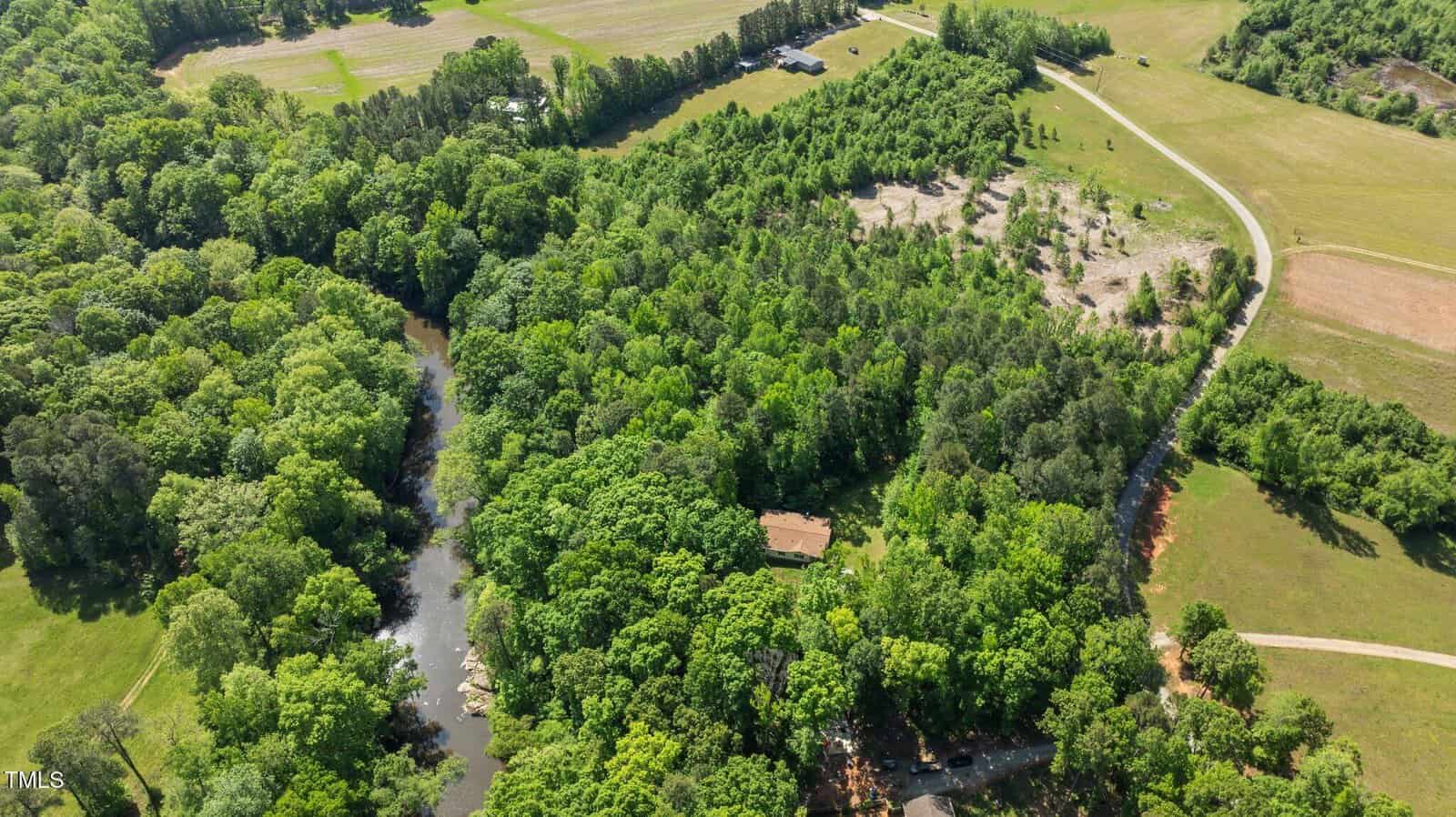 Aerial view of a rural landscape featuring dense green forest, a winding river, a few open fields, and a dirt road with a house partially visible among the trees.