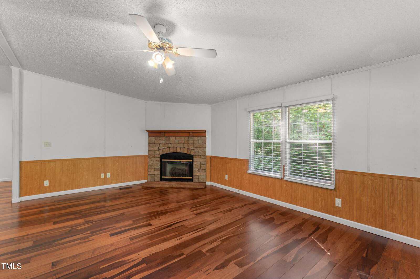 A living room with wood flooring, white walls with wood paneling, a ceiling fan with lights, a stone fireplace in the corner, and two large windows with blinds letting in natural light.
