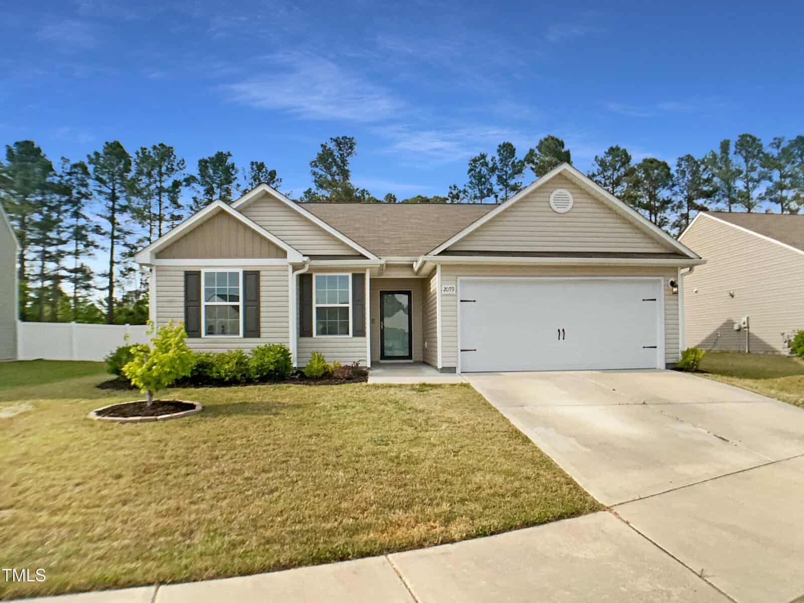 Charming single-story house at 2079 Alderman Way, Creedmoor, with tan siding, white trim, and a two-car garage. A small front lawn features a young tree and concrete driveway, set against trees and blue sky.