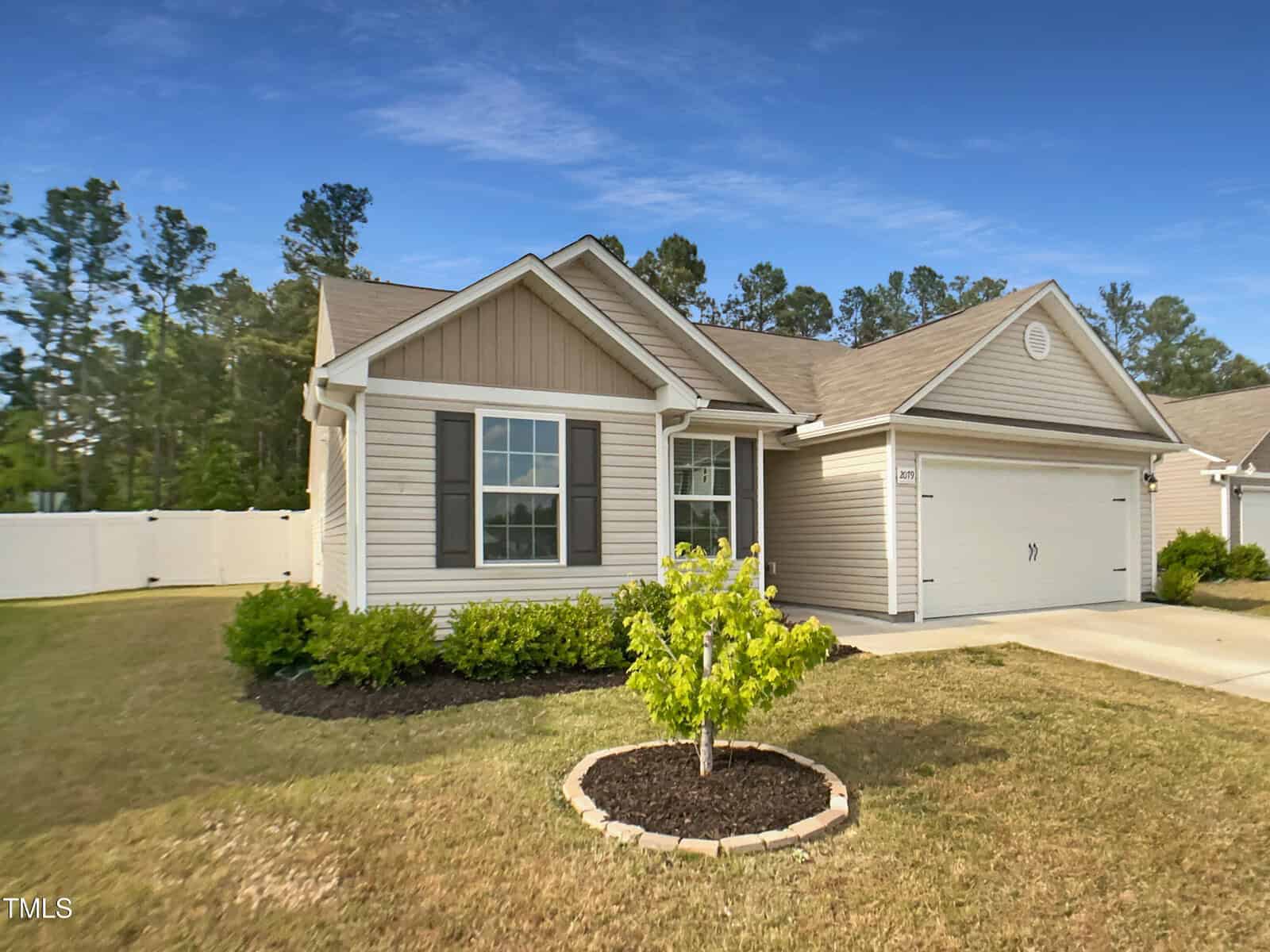 A single-story suburban house with tan siding, white trim, and a double garage. There's a small tree in a mulched circle in the front yard, neatly trimmed bushes, and a white privacy fence along the side.