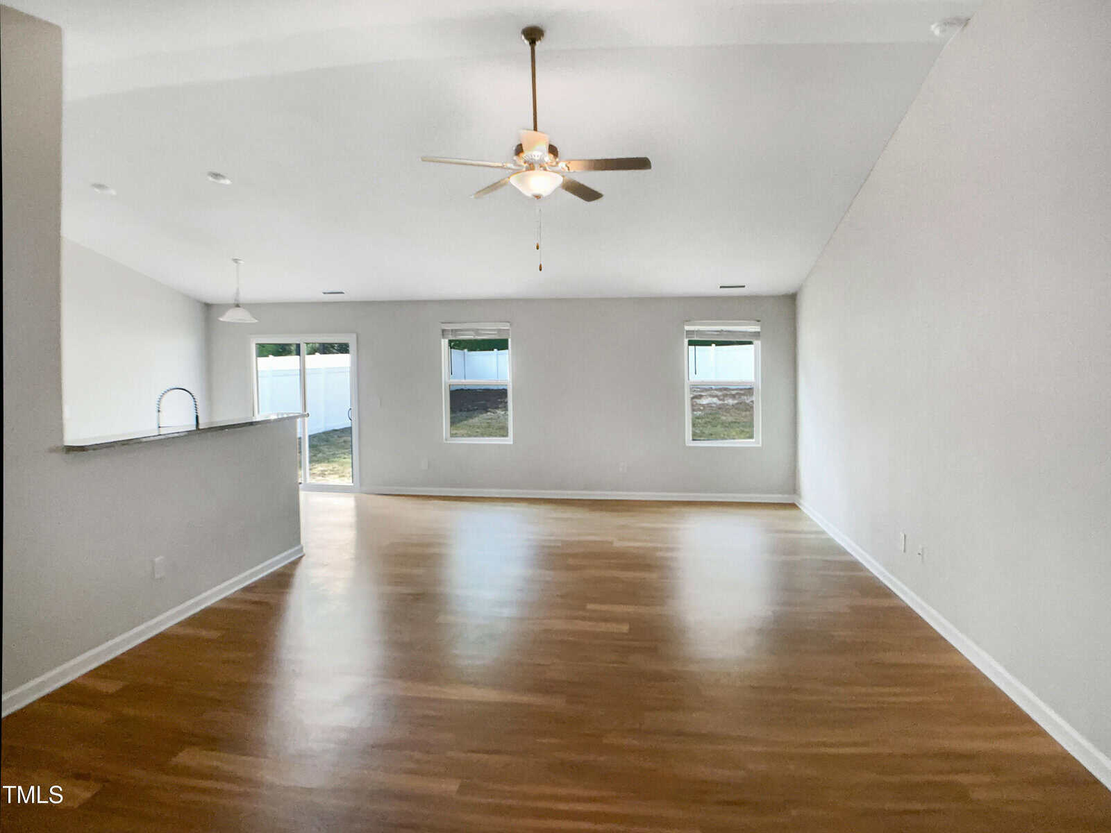 A spacious, empty room with light wood flooring, white walls, two windows, a glass door leading outside, a ceiling fan with lights, and a kitchen area with a counter and sink visible on the left.