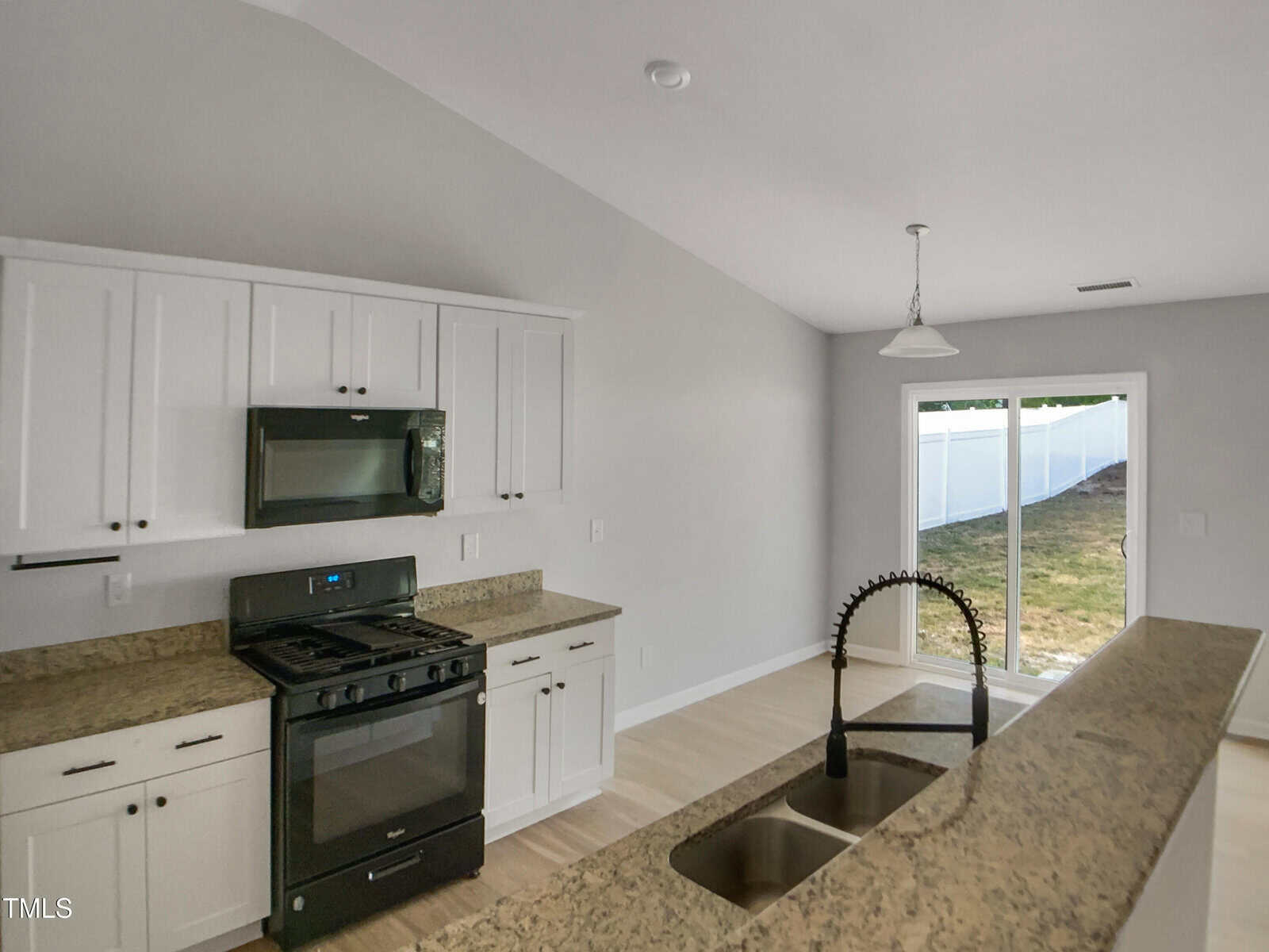 A modern kitchen with white cabinets, granite countertops, a black gas stove and microwave, a double sink with a gooseneck faucet, and a sliding glass door leading to a fenced backyard.