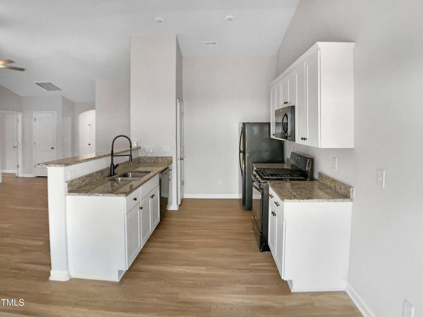 A modern kitchen with white cabinets, stainless steel appliances, granite countertops, a double sink with a black faucet, and light wood flooring. The kitchen is open to an adjacent room.