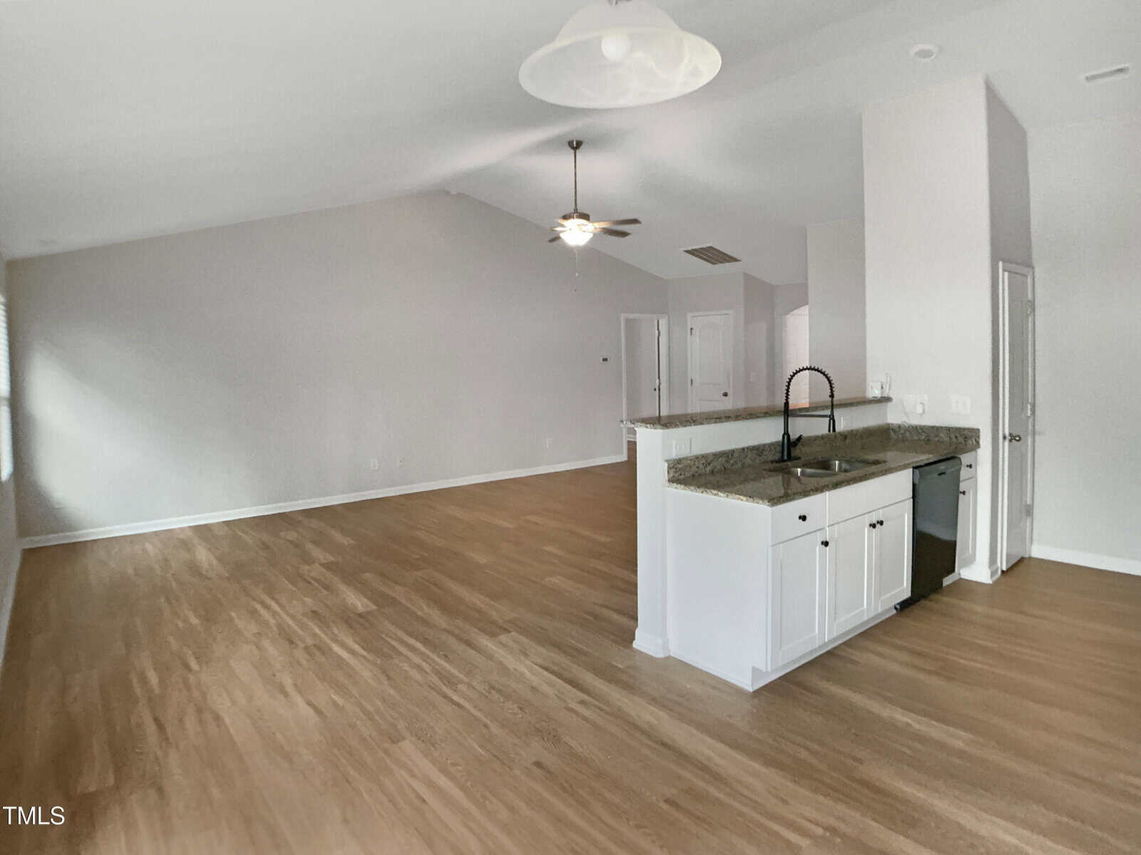 Open concept living area with light wood flooring, white walls, and a ceiling fan. The kitchen features white cabinets, granite countertops, a black faucet, and stainless steel appliances. Natural light enters from the left.