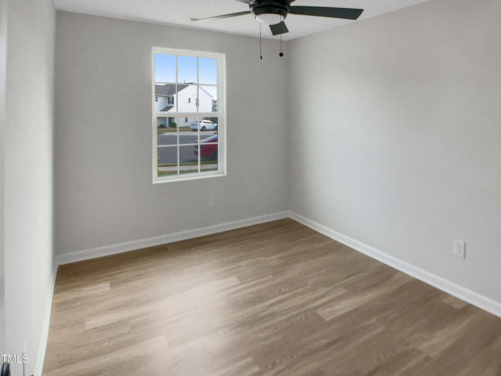 A small, empty room with light-colored walls, light wood flooring, a window showing houses outside, a ceiling fan with lights, and white trim along the floor and window.