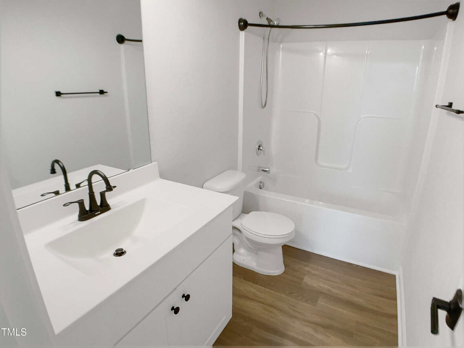 A bathroom with a white vanity and black faucet, a white toilet, a white bathtub with a shower, black fixtures, and wood-style flooring. The walls are painted white.