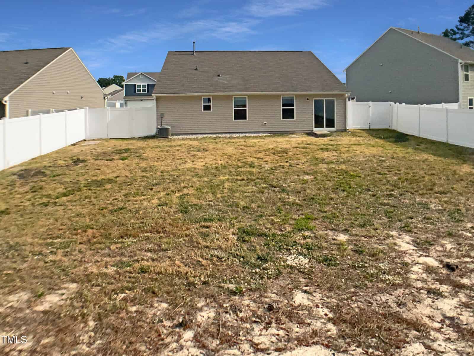 A fenced backyard with patchy grass and some bare spots, behind a single-story beige house with a gable roof, three windows, a sliding glass door, and neighboring houses on both sides.