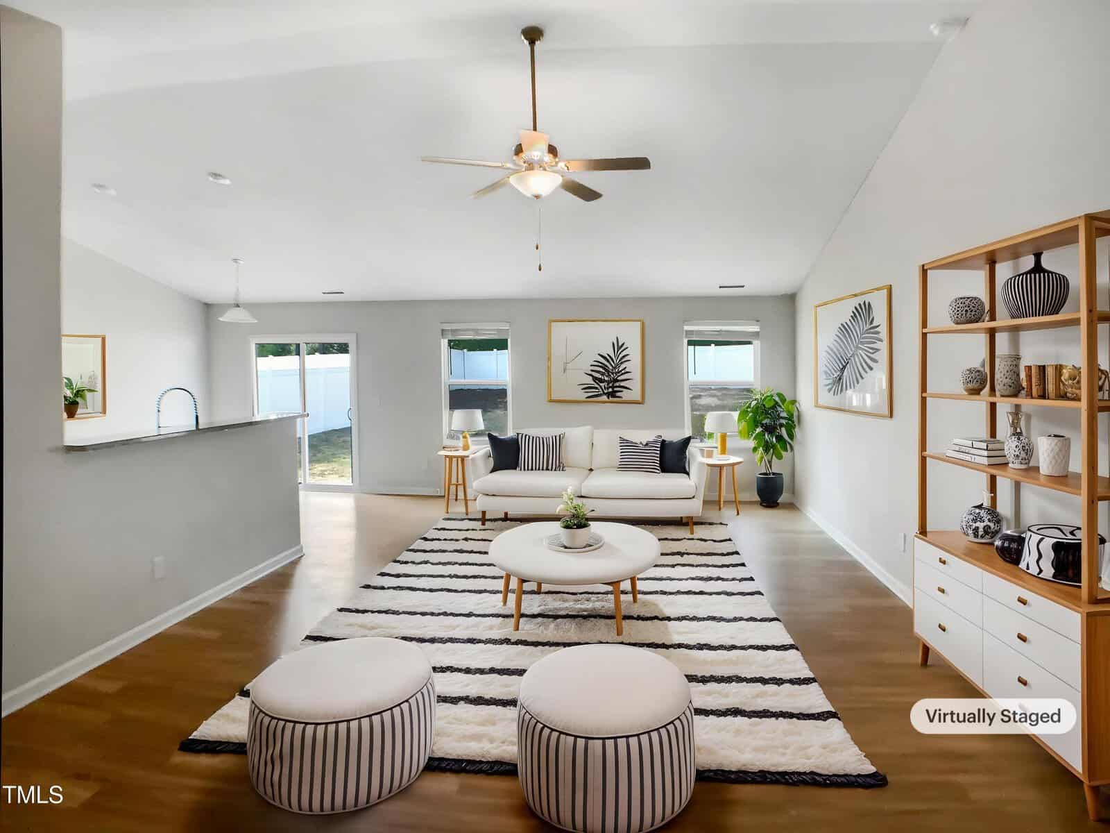 A modern living room with light walls, striped poufs, a round coffee table, a sofa, and open shelving with decor. A black and white rug covers the floor, and framed art hangs on the back wall. A ceiling fan is overhead.