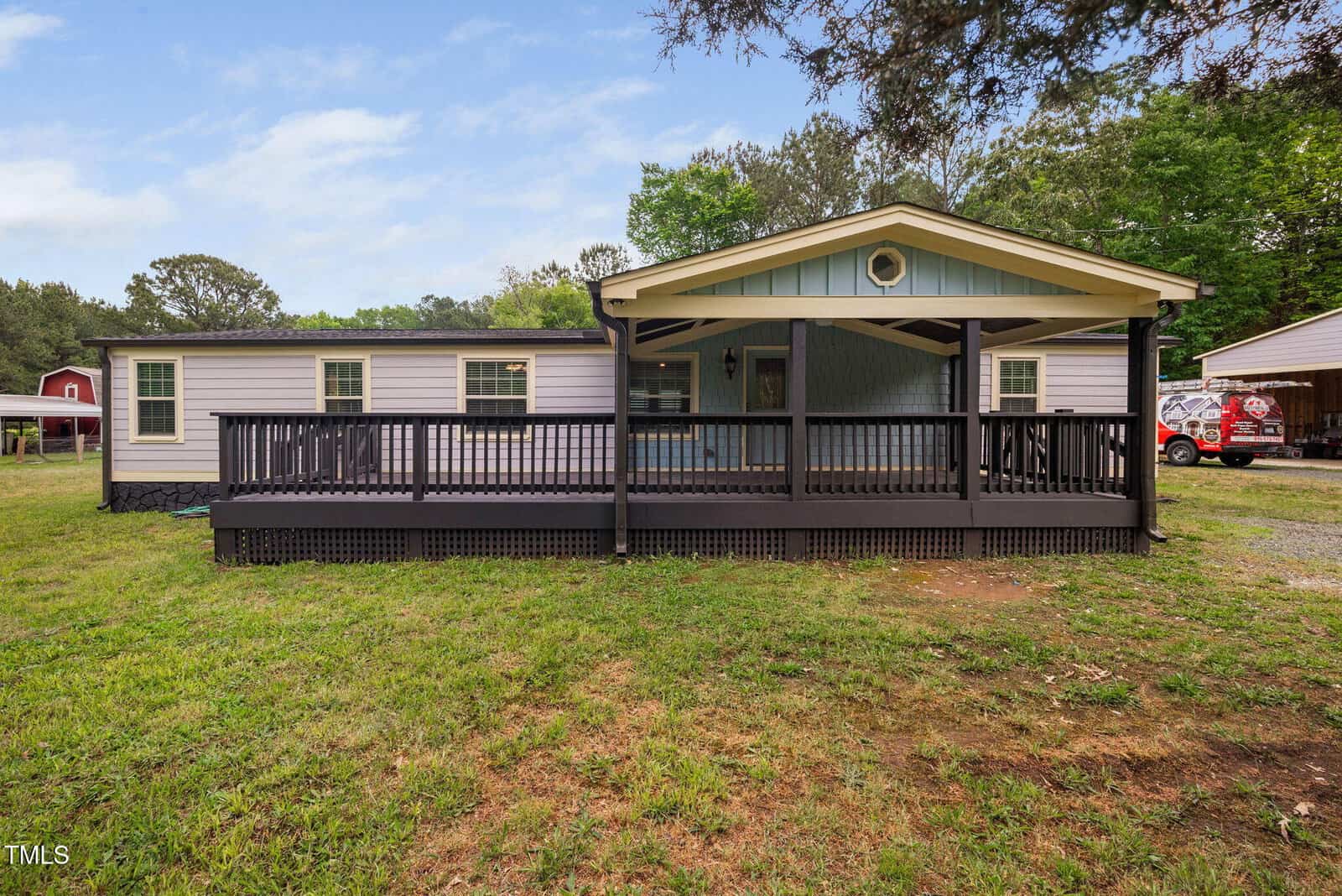 Single-story manufactured home with a wide, covered front porch and dark railing, set on a grassy lot with trees and a clear sky in the background. A detached red barn is visible to the left.