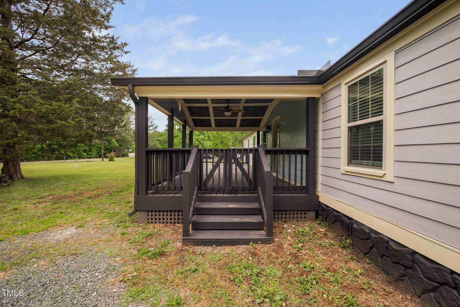 A small wooden porch with black railings and steps leads to the side entrance of a gray house. The porch is covered, and there are trees and grass in the surrounding yard.