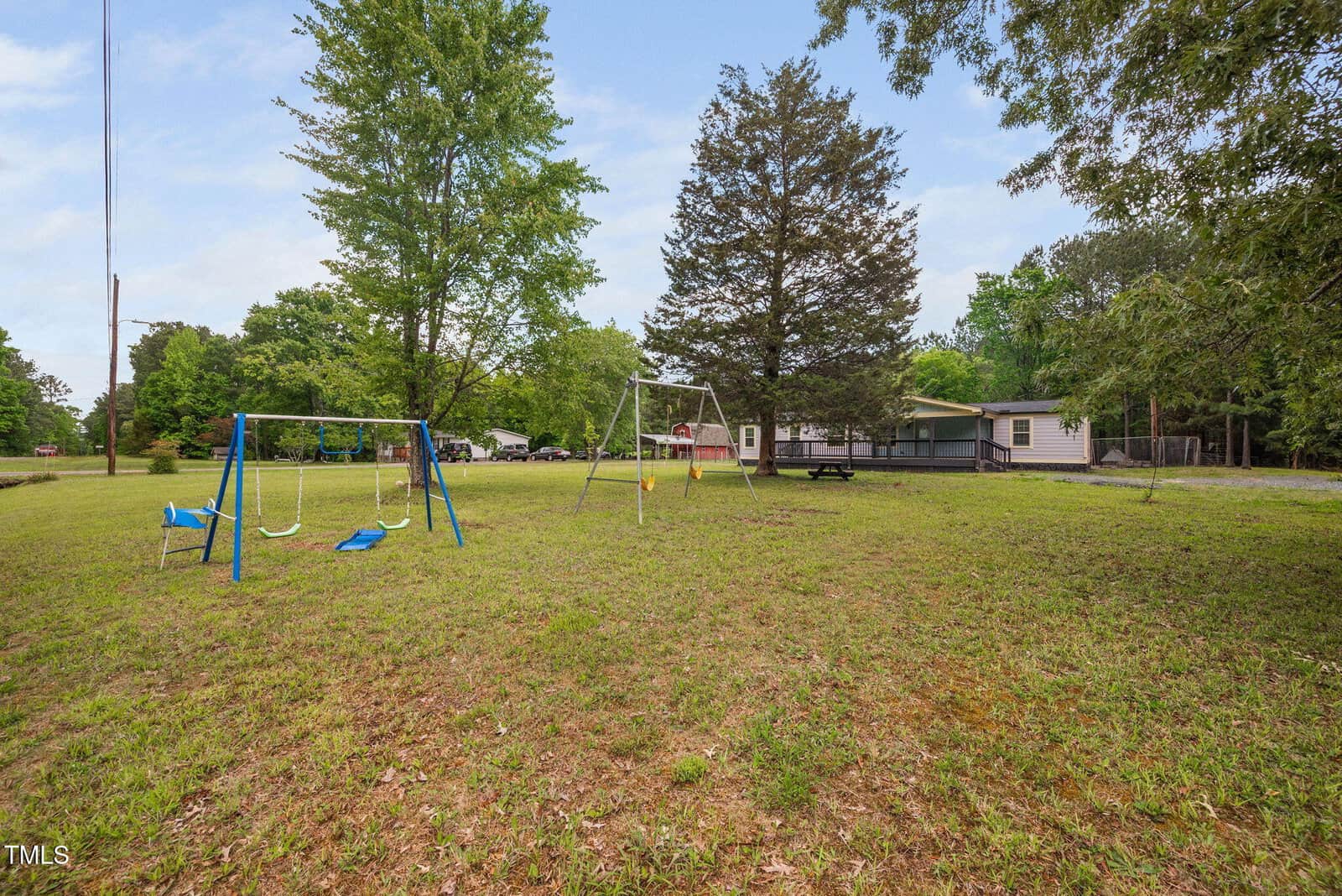 A grassy yard with two swing sets, surrounded by trees. In the background, there is a single-story house with a covered porch. The sky is mostly clear.